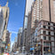 A city street lined with tall buildings under a clear blue sky, with traffic and pedestrians visible.