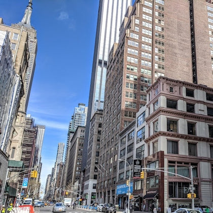A city street lined with tall buildings under a clear blue sky, with traffic and pedestrians visible.