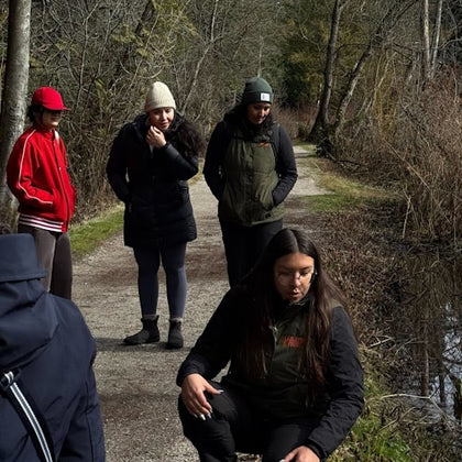 A group of people on a forest path, some standing and one crouching near the ground, with trees and bushes in the background.