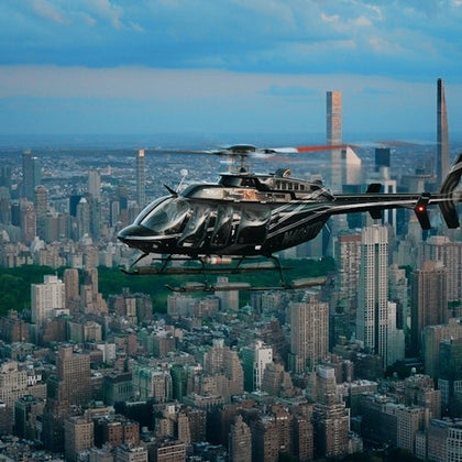 A black helicopter flying over a densely packed city skyline with high-rise buildings and greenery under a cloudy sky.
