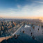 People on a rooftop observation deck overlooking a city skyline at sunset, with glass railings and wide concrete steps.