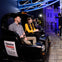 People sit in a black ride car at an indoor attraction with a sign that reads "TheatrePeople.com." The area is dimly lit with blue accents.