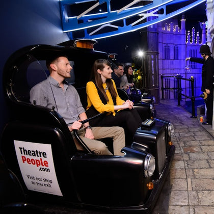 People sit in a black ride car at an indoor attraction with a sign that reads "TheatrePeople.com." The area is dimly lit with blue accents.