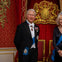 A man and a woman in formal attire stand in front of a red and gold ornate backdrop with two red and gold chairs.