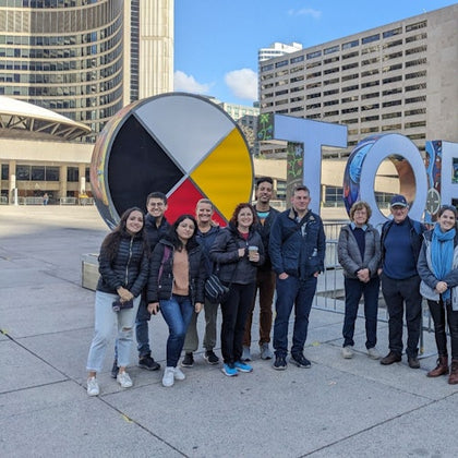 A group of people stand in front of a large sign with colorful letters in an urban plaza with modern buildings.
