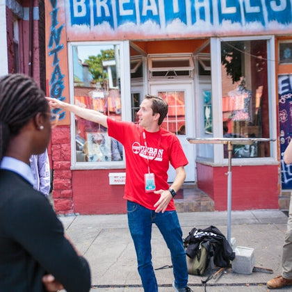 A man in a red shirt points while standing in front of a vintage shop; two others look on.