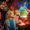 A group of children and an adult interact with a person in a costume near a glowing crystal ball displaying an animated green face.