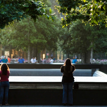 Two people stand at the edge of a large memorial fountain surrounded by trees, with more people visible in the background.