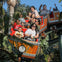 A group of people riding a wooden-themed roller coaster surrounded by tropical plants, smiling and raising their hands.