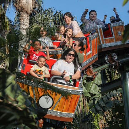 A group of people riding a wooden-themed roller coaster surrounded by tropical plants, smiling and raising their hands.