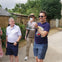 A group of people standing outdoors on a path with greenery and stone walls in the background, one holding a picture.