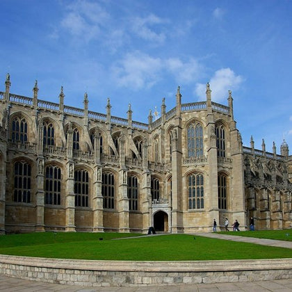 A historic stone building with large Gothic-style windows and spires, surrounded by a green lawn under a blue sky.