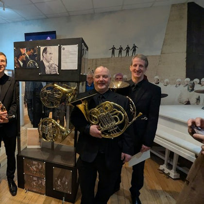 Three men in formal attire holding French horns stand beside a display case with pictures and instruments in a room with white walls.