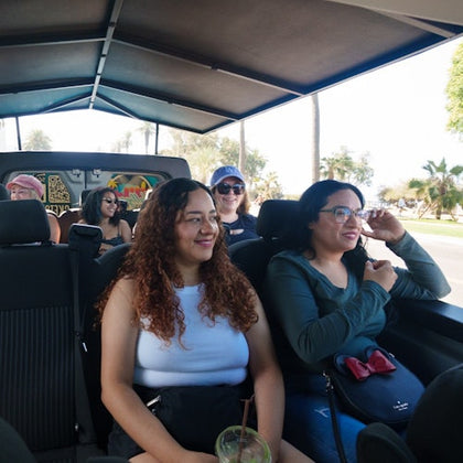 A group of people sitting in an open vehicle under a canopy, smiling. Palm trees and a sunny background are visible.