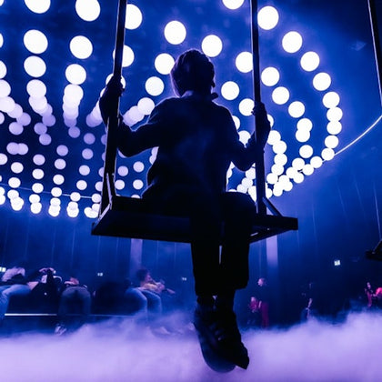 A silhouetted person sits on a swing against a background of glowing circular lights and fog in a dark setting.