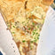 A slice of pizza with a sesame seed crust, white sauce, cheese, parsley garnish, and toppings, served on a paper plate.