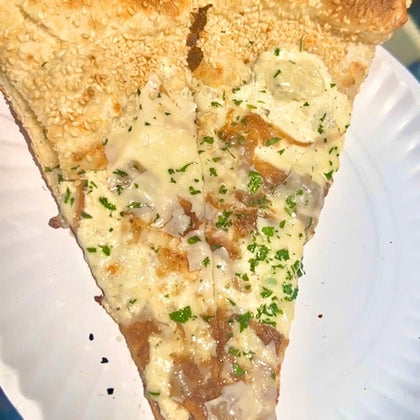 A slice of pizza with a sesame seed crust, white sauce, cheese, parsley garnish, and toppings, served on a paper plate.