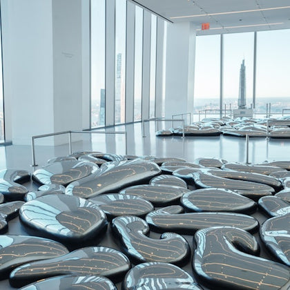 A modern gallery space with large, reflective, organic-shaped sculptures on the floor. Tall windows offer a city skyline view.