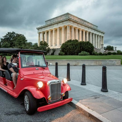 A red vintage-style tour vehicle with passengers in front of the Lincoln Memorial on a cloudy day.