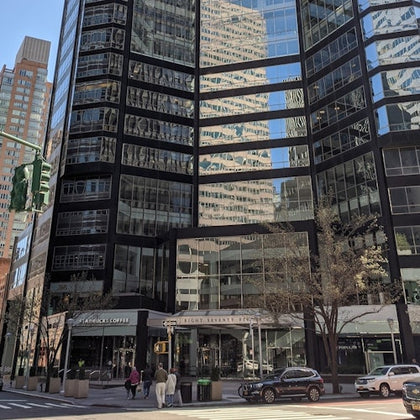 A modern glass office building reflecting city surroundings with pedestrians and vehicles in front on a sunny day.