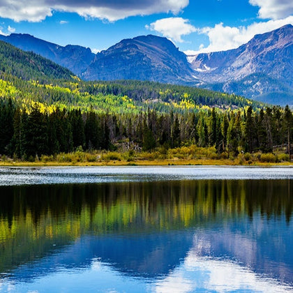 A serene mountain landscape with a forest and a lake, reflecting the trees and sky, under a partly cloudy blue sky.