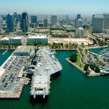 Aerial view of a naval ship docked at a harbor with a city skyline in the background and a marina visible on the right.