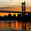 A suspension bridge silhouetted against a vibrant orange and yellow sunset with a city skyline and water below.