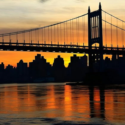 A suspension bridge silhouetted against a vibrant orange and yellow sunset with a city skyline and water below.