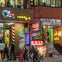 A busy street scene with various illuminated signs for restaurants and shops, people walking, and a cyclist riding by.