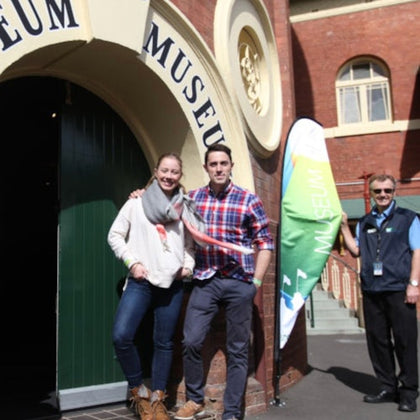 Two people stand smiling in front of a museum entrance, with a staff member and a flag nearby on a sunny day.