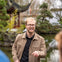 A man in a tan jacket and glasses standing in a garden with a pond and traditional-style pavilion in the background.