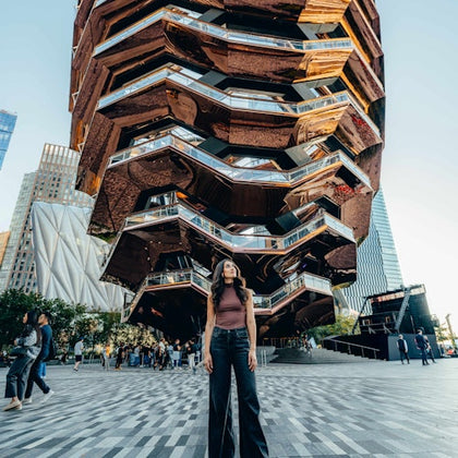 Woman standing in front of the Vessel, a honeycomb-like structure, with surrounding people and buildings in the background.