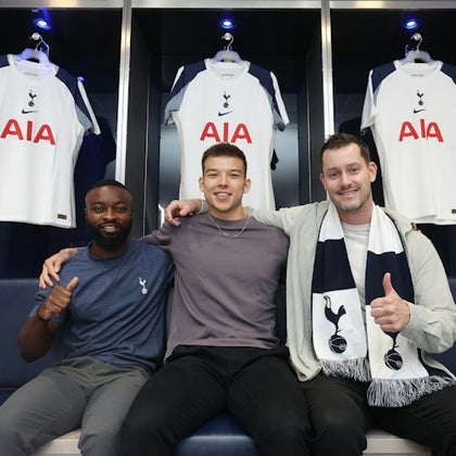 Three men sit on a bench in a locker room, each giving a thumbs-up, with Tottenham Hotspur football jerseys hanging behind them.