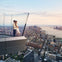 A couple standing on a glass balcony overlooking a city skyline at sunset, with a river in the background.