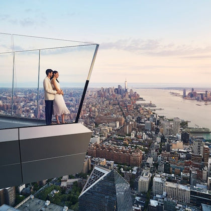 A couple standing on a glass balcony overlooking a city skyline at sunset, with a river in the background.