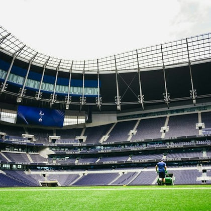 A person operates a lawn mower on a bright green field inside a large, empty stadium with tiered seating and a partial roof.