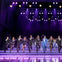 Performers in suits and hats dancing in unison on a stage under purple lighting, with a live band in the background.