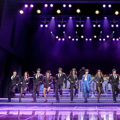 Performers in suits and hats dancing in unison on a stage under purple lighting, with a live band in the background.