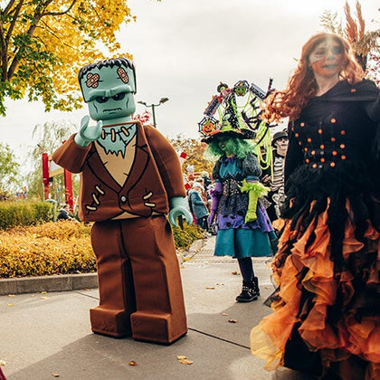 People dressed in Halloween-themed costumes, including a Frankenstein-like Lego figure, walking on a park pathway with autumn trees.