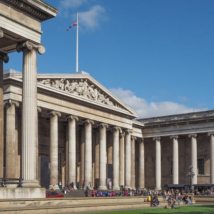A neoclassical building with columns and a triangular pediment, featuring people gathered in the foreground under a blue sky.