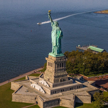Aerial view of the Statue of Liberty on Liberty Island surrounded by water, with a boat in the distance and greenery nearby.