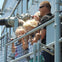 A group of people, including two children, stands on a metal deck near railings, looking out through binoculars on a sunny day.