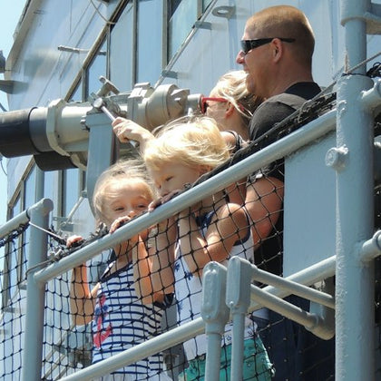 A group of people, including two children, stands on a metal deck near railings, looking out through binoculars on a sunny day.