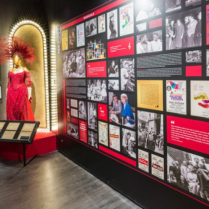 A museum exhibit featuring a red sequined dress with feathers on a mannequin and a wall of historical photos and text displays.
