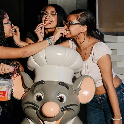 Three people smiling and interacting with a cartoon chef rat statue, in an indoor setting.