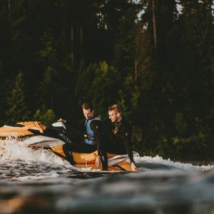 Two people in wetsuits riding an orange jet ski on water, with a forested background.