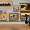 Two people view six landscape paintings in a gallery, arranged in gold frames on a neutral-colored wall.