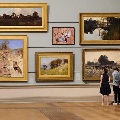 Two people view six landscape paintings in a gallery, arranged in gold frames on a neutral-colored wall.