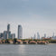 View of a city skyline with modern skyscrapers behind a waterfront area featuring low-rise buildings with red roofs and greenery.