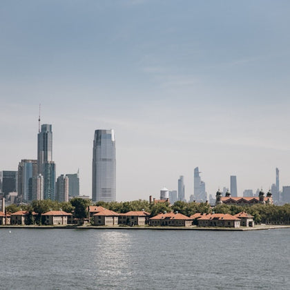 View of a city skyline with modern skyscrapers behind a waterfront area featuring low-rise buildings with red roofs and greenery.